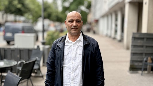 Ein Mann mit schwarzer Jacke und weißem Hemd bekleidet steht auf einem Bürgersteig, links stehen drei Stühle und ein runder Tisch.