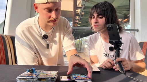 Zwei Männer schauen sich Pokemon Karten an. Die Pokemon Karten liegen auf einem Tisch. Es steht ein Handy auf einem Stativ daneben.