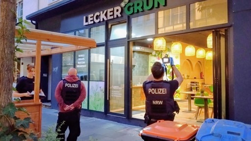 Zwei Polizeibeamte stehen vor dem Ladenlokal "LECKER GRÜN", einer fotografiert den Innenraum, links im Bild ist eine Kellnerin mit Tablett.