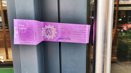 Ein Polizeisiegel der Polizei Wuppertal klebt an einem grauen Türrahmen, in der Mitte ist das Landeswappen von NRW.