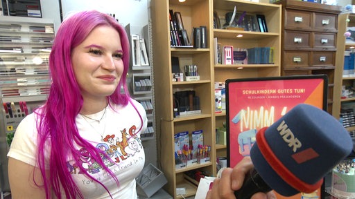 Eine junge Frau mit rosa Haaren wird in einem Schreibwarengeschäft interviewt