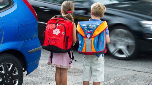 Zwei Kinder stehen mit Schulrucksack zwischen Autos und warten an einer Straße