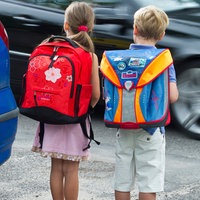 Zwei Kinder stehen mit Schulrucksack zwischen Autos und warten an einer Straße