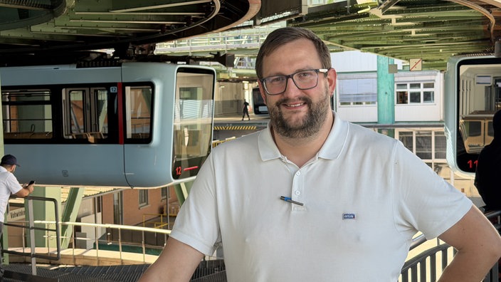 Ein Mann steht vor einer Schwebebahn und lächelt in die Kamera.