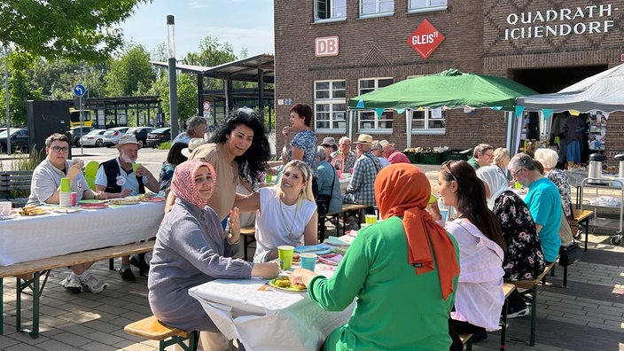 zu sehen sind viele Menschen, die sich auf Bierbänken zusammengesetzt haben. Sie essen und trinken zusammen am Bergheimer Bahnhof