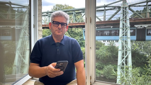 Ein Mann mit Brille sitzt an einem geöffneten Fenster, hinter ihm fährt die Wuppertaler Schwebebahn vorbei