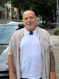 Ein Mann mit grauem Bart steht vor einem geparkten Auto
