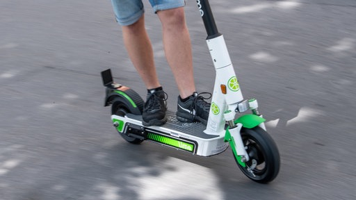 Ein Mann in kurzen Jeans und mit schwarzen Sportschuhen steht auf einem E-Roler und fährt, zu sehen ist er nur knieabwärts