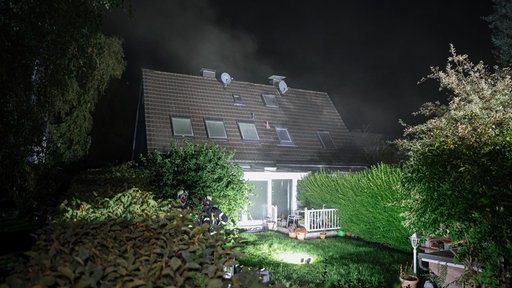 Nächtliche Außenansicht eines Wohnhauses mit Garten, beleuchteter Fassade und leichtem Qualm über dem Dach.