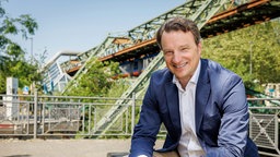 Dr. Hagen W. Lippe-Weißenfeld sitzt vor der Schwebebahnbrücke