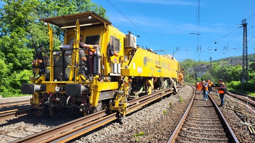 Ein große, gelbe Bahn. Auf den Gleisen daneben befinden sich Menschen in Warnwesten und Helmen.