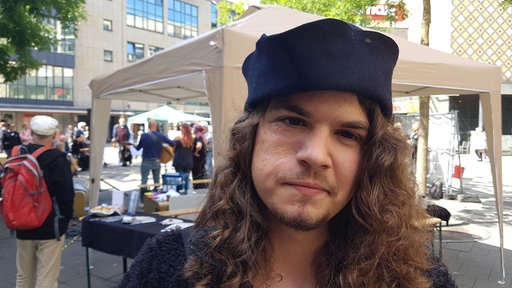 Ein Mann mit langen gelockten braunen Haaren steht in einer Fußgängerzone, auf dem Kopf trägt er eine schwarze Mütze
