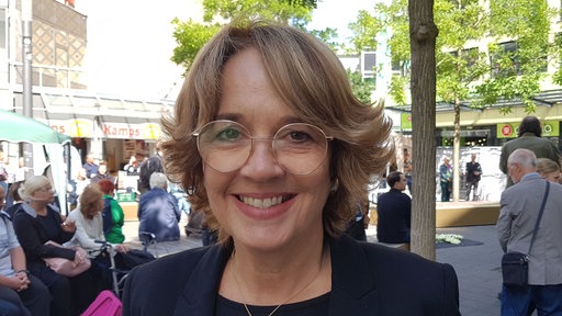 Eine Frau mit Brille steht in einer Fußgängerzone und lächelt 