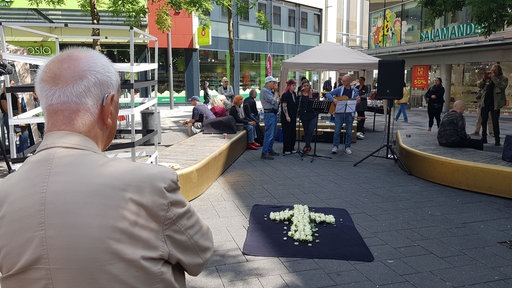 Ein weisses Blumenkreuz auf dem Boden in einer Fußgängerzone, im Hintergrund stehen Passanten