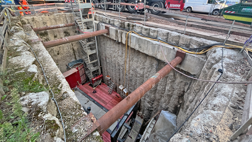 Eine Baugrube neben einer Straße. An der Seite führt eine Bautreppe nach unten. Oben stützen drei Metallstützen die Seiten ab.