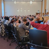 Man sieht einen Stadtratsaal gefüllt mit Menschen. Viele heben die Hand.
