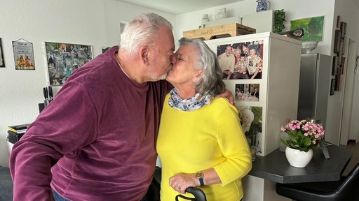 Ein älteres Paar gibt sie einen Kuss. Der Mann hat seinen Arm um die Frau gelegt. Im Hintergrund sind viele Bilder zu sehen.