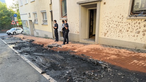 Drei Polizisten vor einer Hauswand mit verbanntem Asphalt vor ihnen