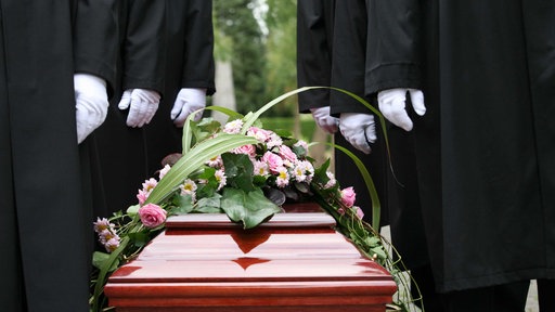 Ein brauner Sarg mit Pink-weißen Blumen drauf, daneben stehen Männer in schwarzen Anzügen und weißen Handschuhen