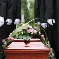 Ein brauner Sarg mit Pink-weißen Blumen drauf, daneben stehen Männer in schwarzen Anzügen und weißen Handschuhen