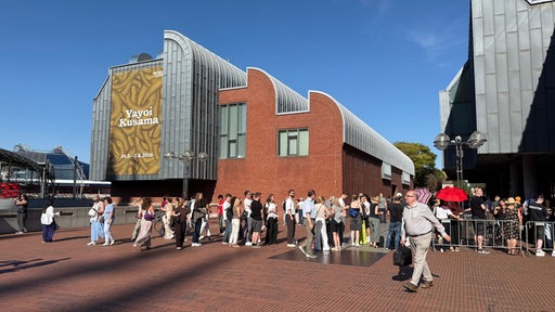 Menschenmasse vor einem Gebäude auf der ein Kusama Plakat hängt