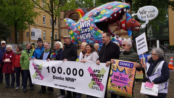 Die Initiatoren der Unterschriftenaktion halten ein Banner in den Händen: 100.000 Unterschriften. Im Hintergrund eine Jacques Tilly Figur. Ein großer bunter Fisch mit der Aufschrift "wir sind mehr" frisst einen kleineren AfD Fisch.