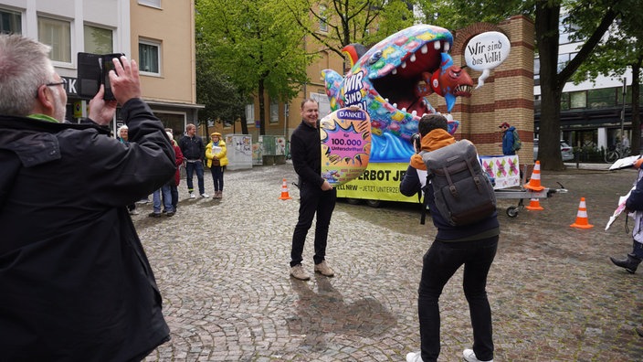 Der Erfolg von 100.000 Unterschriften für ein AFD-Verbot wird auf Fotos festgehalten. Jacques Tilly posiert vor seinem Karnevalswagen.