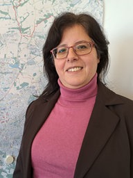 Eine Frau mit dunkelbraunen mittellangen Haaren lächelt in die Kamera. Sie steht vor einer Wand, an der eine Karte von Bielefeld hängt.