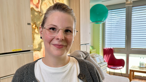 Eine Frau mit Zopf und Brille steht in einem Kreißsaal und blickt in die Kamera. 