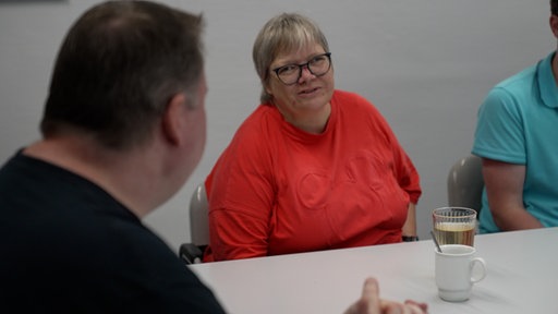 Eine Frau im roten Shirt sitzt an einem Gruppentisch und schaut zu einem Mann im schwarzen Shirt.