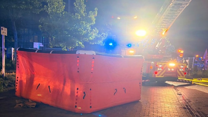 Ein großes, rotes Sprungpolster liegt auf der Straße, im Hintergrund ein Einsatzwagen der Feuerwehr mit Drehleiter.