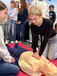 Eine junge Frau mit blonden Haaren, schwarzem Oberteil und helblauer Jeans kniet auf einer roten Matte auf dem Boden. Sie presst mit beiden Händen auf den Oberkörper einer Reanimationspuppe. Um Sie herum sind viele andere junge Menschen zu sehen.