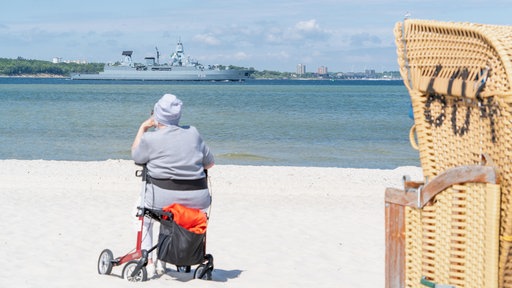 Eine alte Frau sitzt auf einem Rollator am Strand.