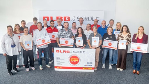 Mehrere Menschen stehen für Bild in Reihen und halten Urkunden auf denen "DLRG & Schule" steht.