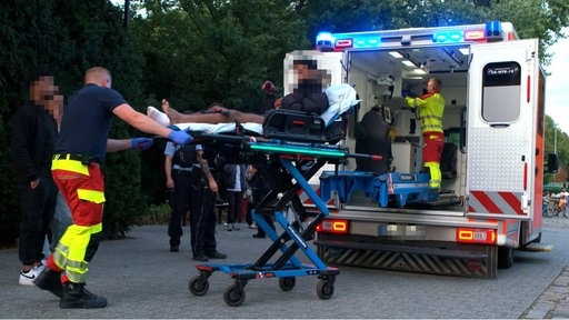 Ein Mann wird auf einer Trage in einen Krankenwagen geschoben. Mehrere Personen stehen um ihn.
