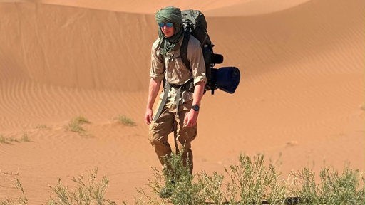 Tim Jambor läuft mit Sonnenbrille und Kopftuch durch die Sahara. Im Hintergrund ist eine Düne zu sehen. 