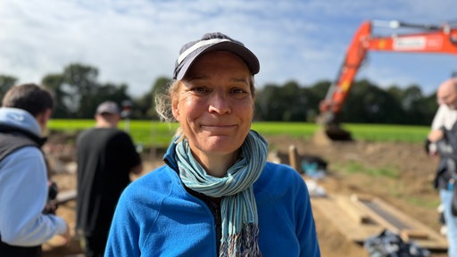 Eine Frau mit blauem Pullover und grauer Kappe steht vor einer Ausgrabungsstelle und lächelt in die Kamera. 
