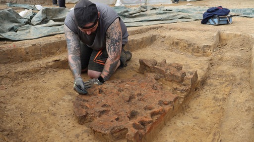 Ein Mann hockt in einer Grabungsfläche auf braunem Boden und bearbeitet mit dem Pinsel einen Fund einer archäologischen Ausgrabung. 
