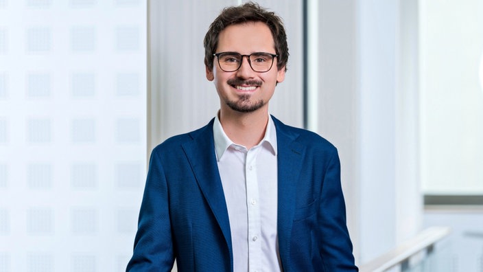 Ein Mann mit braunen Haaren, Bart und Brille lächelt im blauen Anzug in die Kamera.