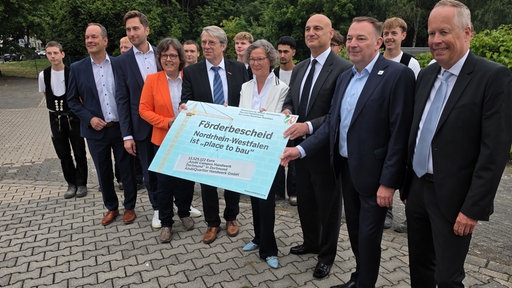 Mehrer Menschen in schicker Kleidung halten ein großes Plakat mit der Aufschrift "Förderbescheid".