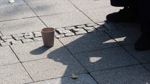 Vor dem Schatten einer bettelnden Person steht ein Becher auf dem Boden.