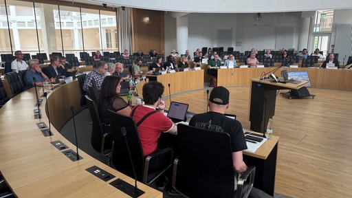 Viele Menschen sitzen im Saal auf schwarzen Lederstühlen.