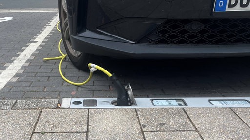 Im Bordstein ist eine Steckdosenleiste eingelassen, daneben steht ein dunkles Auto, das mit einem gelben Stromkabel mit der Steckdose verbunden ist