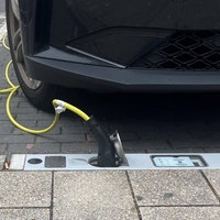 Im Bordstein ist eine Steckdosenleiste eingelassen, daneben steht ein dunkles Auto, das mit einem gelben Stromkabel mit der Steckdose verbunden ist