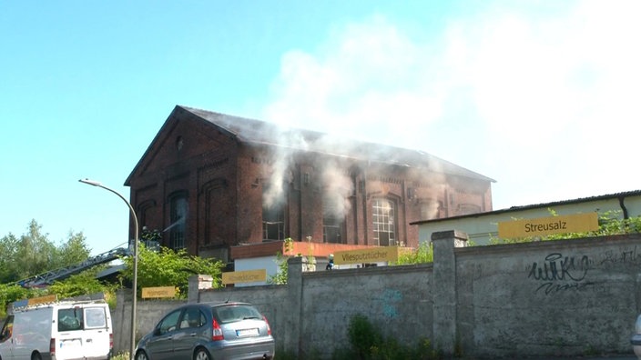 Ein großes aus Backsteinen bestehendes Gebäude brennt. Rauch qualmt aus den großen Fenstern.