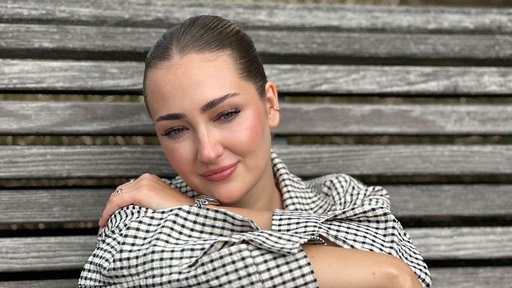 Betroffene Maxine Reuker sitzt die Beine umarmend auf einer Bank.