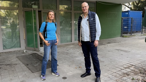 Frau mit türkisem Oberteil und Mann in Weste stehen vor einer großen Fenstertür, dem Ladenlokal des Grünen Salon.