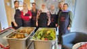 Sieben Frauen in Schürzen mit der Aufschrift "Kochpioniere", davon große Behälter mit Essen.