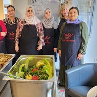Sieben Frauen in Schürzen mit der Aufschrift "Kochpioniere", davon große Behälter mit Essen.