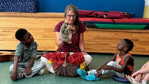 Petra Kern sitzt mit einer Igel-Handpuppe zwischen zwei Kindern, die sie interessiert anschauen.